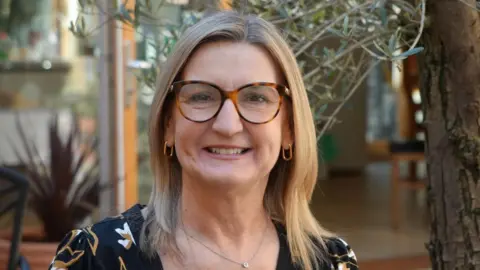 Shaun Whitmore/BBC Jane Featherstone has blonde, shoulder-length hair and she is wearing tortoise shell glasses and a black top with gold and white detailing.