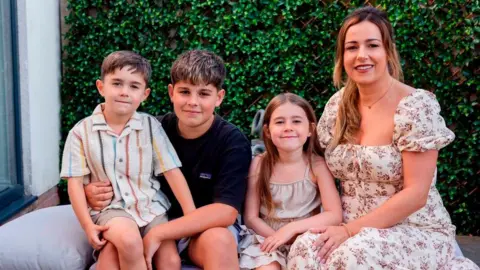 Graham Davies Photography Three children sat next to their mum on a sofa. There is a hedge behind them. Jodanna wears a white and pink floral dress and has a young girl next to her. An older boy has Hudson on his lap. 