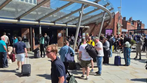 Nick Cosgriff Queues of people outside a brick building with a metal lattice around it. Some pull suitcases.