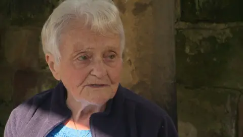 A woman with short white hair, wearing a blue fleece and sitting in front of a stone wall.