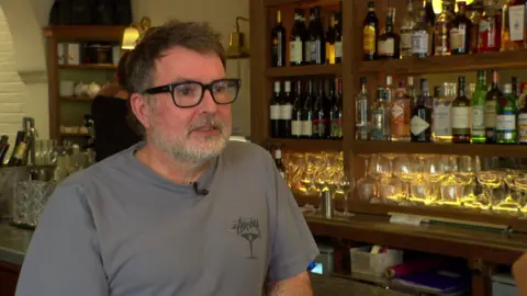 Jamie Younger, a middle aged white man in his pub. He is wearing a grey t shirt and black glasses.