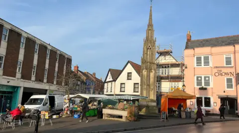 The picture shows Market Cross in Glastonbury.  The picture shows stalls and people walking around. The Crown pub is in the background.