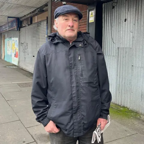 Kevin Shoesmith/BBC News John Holmes carries a rolled-up copy of a newspaper as he stands in Freeman Street, Grimsby, with boarded-up shops behind him. He is wearing a grey jacket and a blue flat cap