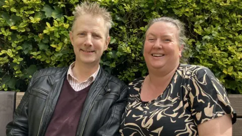 A man wearing a striped top, dark purple jumper and a leather jacket. He has blonde hair and is smiling. A woman wearing a black and light brown dress. Her hair is tied back. She is smiling at the camera. They are both sat on a brown bench. There is a green bush behind them. 