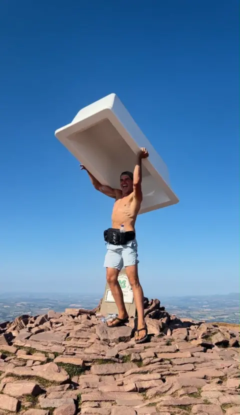 Tom Christopher A shirtless man is lifting a bath tub above his head with his arms while standing on stones and rocks on a mountain summit