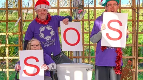 Three people wearing purple T-shirts. One is sitting in a wheelchair, with the other two standing. They are positioned around a toilet in an outdoor setting, holding three placards with the letters "S. O. S."