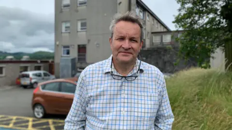 A man in a blue and green checked shirt with grey hair and his glasses around his neck stands in front of a school building
