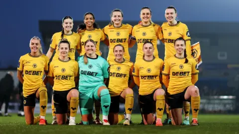 Wolves Women The current Wolves Women team is seen in a stadium. They lined up in rows and wearing orange.