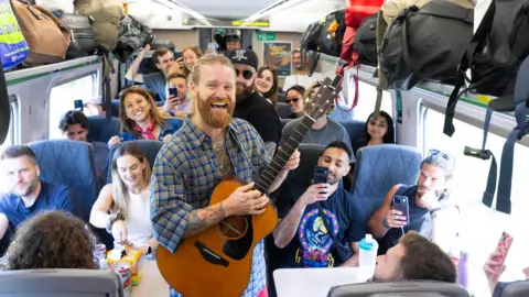 PA Media Sam Ryder holds a guitar as he performs an impromptu gig onboard a train service travelling from Paddington Station to Glastonbury. The train carriage is full of people with several filming on their phones.