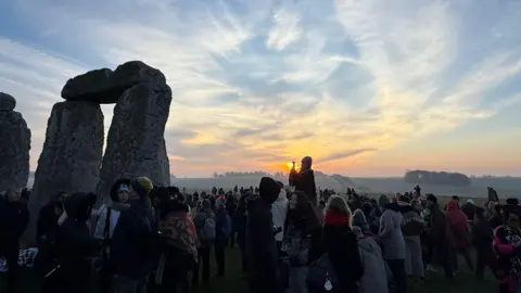 People stood in the sunrise by large stones. One person sits on each another person's shoulders, others stand in groups wrapped up warm in jackets and scarves.