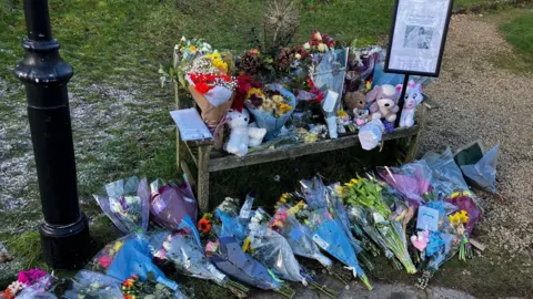 Floral tributes, cuddly toys and messages are laid on a wooden bench and the surrounding floor in a churchyard. There is a very small dusting of snow on the grass beside the bench, and the black pole of a lamp post. A sign bearing a black and white image of a mother and her two children can be seen in a holder.