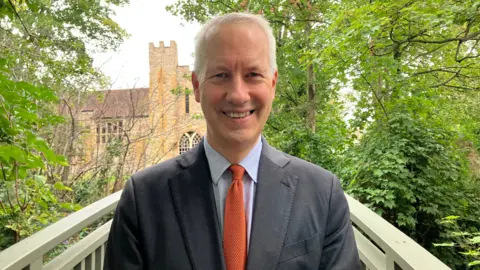A man with a grey suit, blue shirt, and orange tie is looking at the camera. He is surrounded by trees, and behind him in a church.