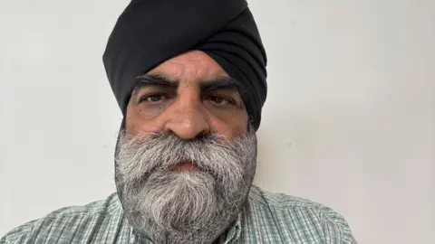 Rajinder Singh Bhogal A man with a white and grey moustache and beard and black turban. He is wearing a green and white checked shirt with red pinstripes.