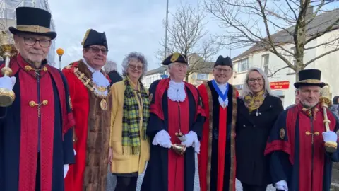 A group of seven people, stand in a line looking at the camera and smiling. Three are wearing long, navy gowns with red detailing on the front, two are wearing long, red gowns with brown fur details and gold chains, and the remaining two are wearing coats - one yellow and one blue - with tartan scarves.