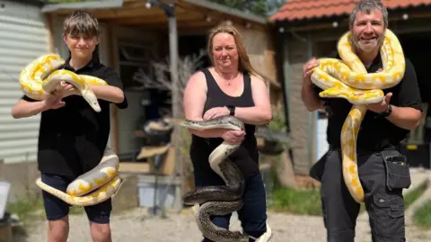 RSG Reptiles A man, a woman and a teenage boy each holding a large snake. One of the snakes is dark brown - the other two have yellow and white markings. They are standing in front of an outbuilding with a tiled roof.