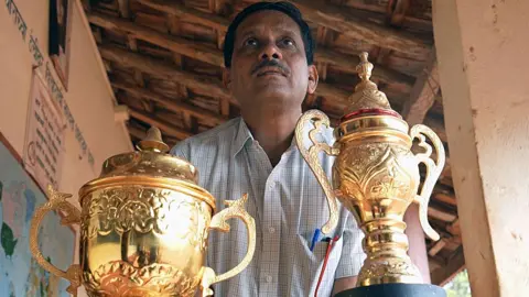 Daji Rajguru in a checked shirt with two large golden trophies in front of him. The trophies are embossed with patterns and are shaped like vases with lids and handles. 