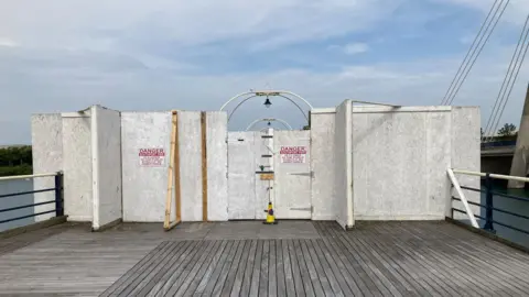 White painted wooden boarding blocks access to the pier. Two signs have anger written on in red writing. and a door in the middle is locked with a padlock.