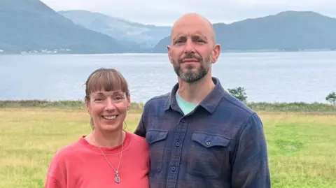 BBC Esther and Nick Cochrane smile as they pose together in front of a stunning loch near their holiday cottage business. Esther wears a red sweatshirt and a pendant, Nick wears a dark blue checked shirt. the landscape is distinctly Scottish.
