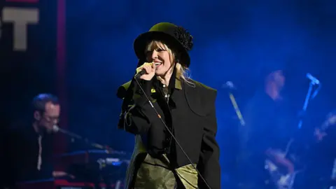 Getty Images Natasha Bedingfield is singing into a microphone on stage. She is wearing a black and green jacket and black hat. She has blonde hair and is smiling. Behind her are a guitarist and a keyboard player.