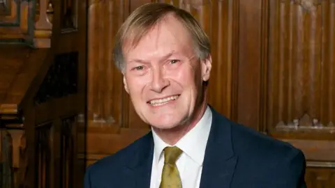 Getty Images Sir David Amess, smiling, wearing a navy suit and olive green tie in front of wooden panels.