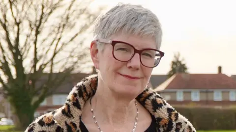 A woman with short cropped grey hair is wearing glasses and a leopard print coat. She is standing outside and smiling