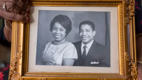 Steve Reeves A woman's hand holds a gilt frame with a black and white photo of two smartly dressed people smiling into the camera