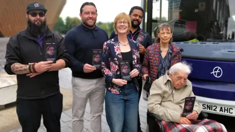 A group of people, including an older man sat in a wheelchair, are stood in front of a bus holding leaflets and smiling.