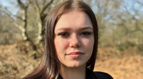 A young woman with long brown hair sits on a park bench on a sunny day. She is wearing a dark green top and black zip-up hoodie, with a small necklace and nose piercing. Bare trees and brown autumn foliage are visible in the blurred background.
