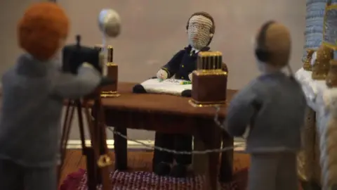 A depiction of a man sitting at a desk, with a camera pointing at him, made out of yarn.