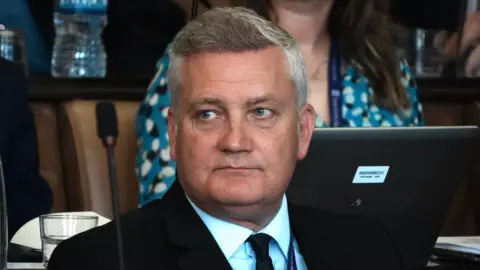 John Fairhall/BBC Russell Quirk is sitting in the council chamber and watching on. He is wearing a suit and has grey hair.