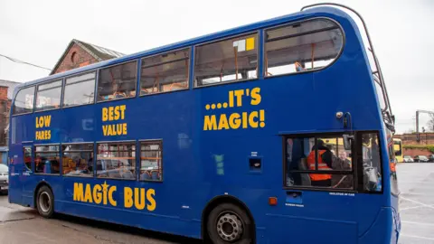 A dark blue double-deck bus parked in a depot on a grey day takes on passengers. The bus has the word's 'Magic Bus' written across in it yellow lettering. 