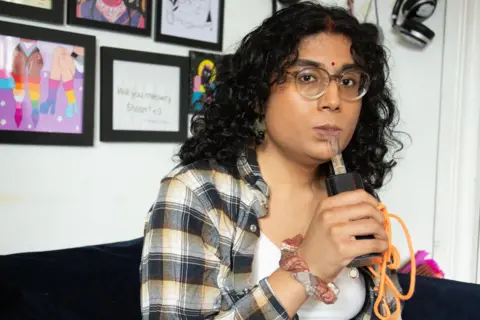 A woman with curly long dark hair and clear rimmed glasses puffs on a large cannabis vaporiser with an orange lead trailing out of it