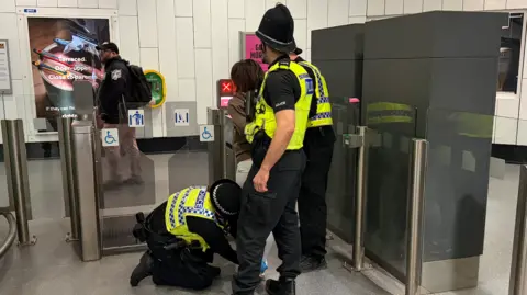 BBC/Jim Scott Three police officers are standing by disabled access entry point at a Metro station. They have detained a woman, who is wearing a brown hoody, and are patting her down to check for weapons or drugs paraphernalia. 