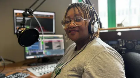 Simon Thake A black woman with light dreadlocks sits in a radio studio wearing headphones
