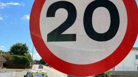 BBC White and red 20mph sign with a backdrop of a residential street