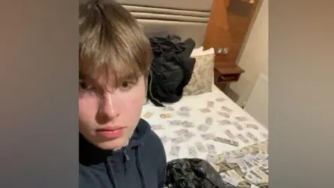 A selfie image of a teenage boy dressed in a black zip up top. He has light brown hair with a long fringe. Behind him s a bed with cash notes laid out and a black bin liner next to them.