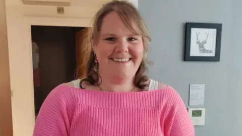 A woman with curly blonde hair smiles at the camera. She wears a pink jumper. She is standing inside.
