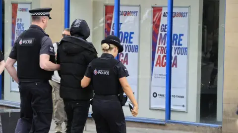 GMP Police take away a person in a black hoodie in a town centre.