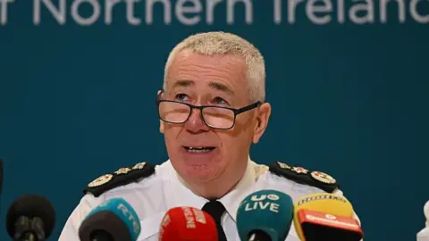 Jon Boutcher speaking into multiple microphones. He is wearing his PSNI uniform - a white shirt with black and gold details, a black tie and is wearing glasses. He has grey hair.