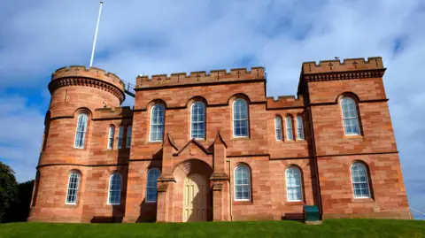 The sandstone castle has towers and battlements. There is a white flag pole.