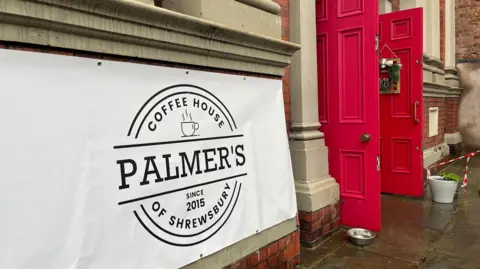 ELLEN KNIGHT/BBC The outside of the Baptist church, which has bright red double doors with green wreaths hanging on them. In the foreground, there's a large white banner that reads: 'Palmers of Shrewsbury, Coffee House, since 2015.' The pavement is wet, suggesting it has recently rained. 
