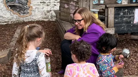 Helen Godwin is wearing a purple blazer and black rimmed glasses. She is smiling at the children who are surrounding her as they play in the nursery playground.