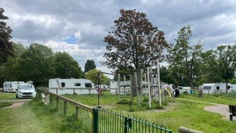 A picture of at least seven caravans parked next to a playpark, alongside a number of other cars and vans. 