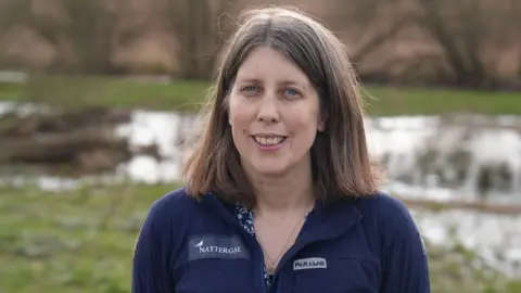 A woman with shoulder-length brown hair stands outdoors in a grassy, waterlogged landscape with soft-focus trees and pools of water behind them. They are wearing a dark blue zip-up jacket with two embroidered logos on the chest, including one reading "Nattergal".