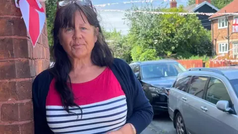 Jonny Humphries/BBC A middle-aged woman with long dark brown hair leans against the brick wall of a house, with England flags visible in the background