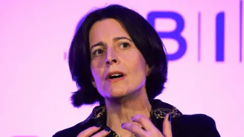 Getty Images A woman with short black hair is speaking on a stage. She is sitting down and is wearing a black jumper. 