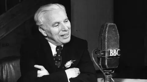 A black and white image of Charlie Chaplin showing him in 1952, talking into a BBC microphone. He is wearing a dark suit, and tie with a flower in his lapel