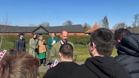 The Duke of Edinburgh speaks to a group of students standing on a field. There are four women standing behind him, one of whom is in a blue uniform