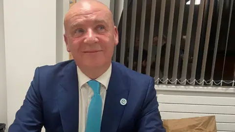 Stuart Keyte, who is wearing a dark blue suit, white short and light blue tie, with a Reform logo pin badge on his jacket. He is bald and is looking slightly off to the side, with a window with blinds behind him. He is smiling.