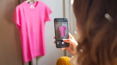 Getty Images A woman taking a photo on her mobile phone of a pink T-shirt hanging up